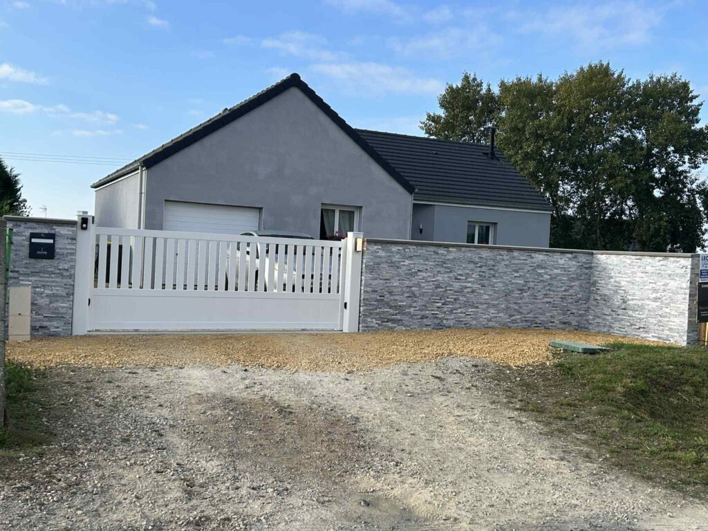 Maison avec mur en pierres grises et portail blanc motorisé après rénovation extérieure dans le Calvados