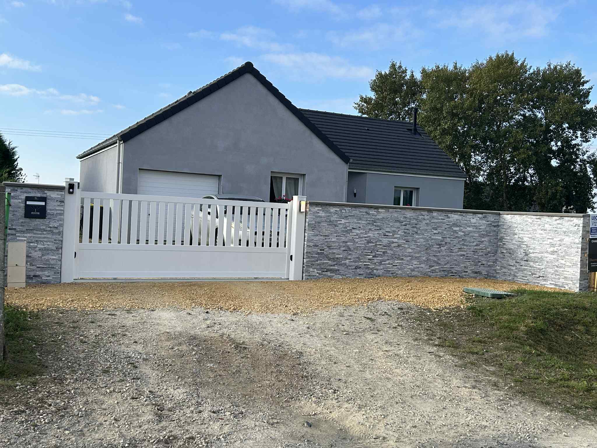 Maison avec mur en pierres grises et portail blanc motorisé après rénovation extérieure dans le Calvados